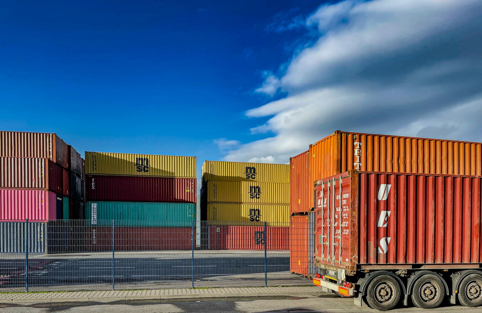 A container loaded on a truck trailer in front of stacked shipping containers